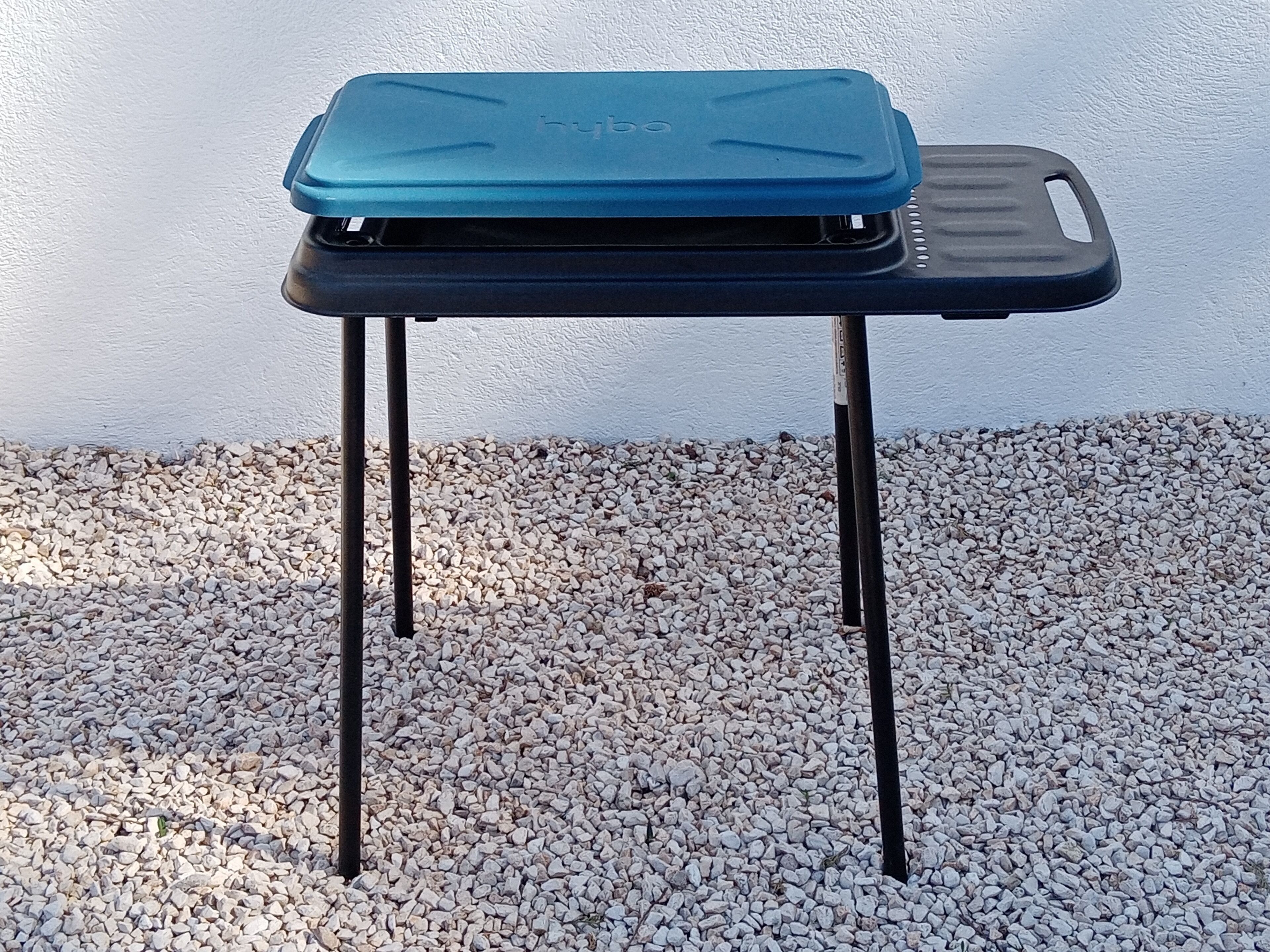 Una mesa plegable con superficie azul y base negra, sobre una superficie de grava frente a una pared blanca.