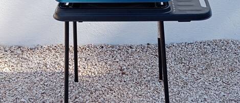 Una mesa plegable con superficie azul y base negra, sobre una superficie de grava frente a una pared blanca.