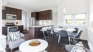 Una cocina moderna con armarios oscuros, un refrigerador de acero inoxidable y un microondas. Hay una zona de comedor con una mesa blanca y sillas grises.