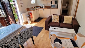 Un espacio habitable compacto con kitchenette, una zona de comedor con mesa y banco, un sofá con cojines estampados, y una puerta que da a otra habitación.