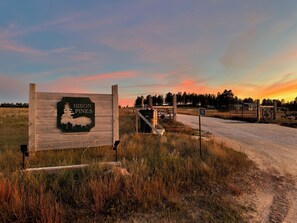 Ein hölzernes Schild mit der Silhouette eines Bären und dem Schriftzug 'Bison Pines' vor einem Sonnenuntergang.