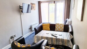 A compact dining area with a table and four chairs, a flat-screen TV mounted on the wall, and a window with curtains. - Stay Norwich Apartments 17Colgate (Norfolk)