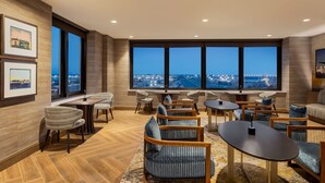 A modern lounge area with a large window offering a city view, wooden flooring, and contemporary furniture. - Marriott New York JFK Airport (Jamaica)