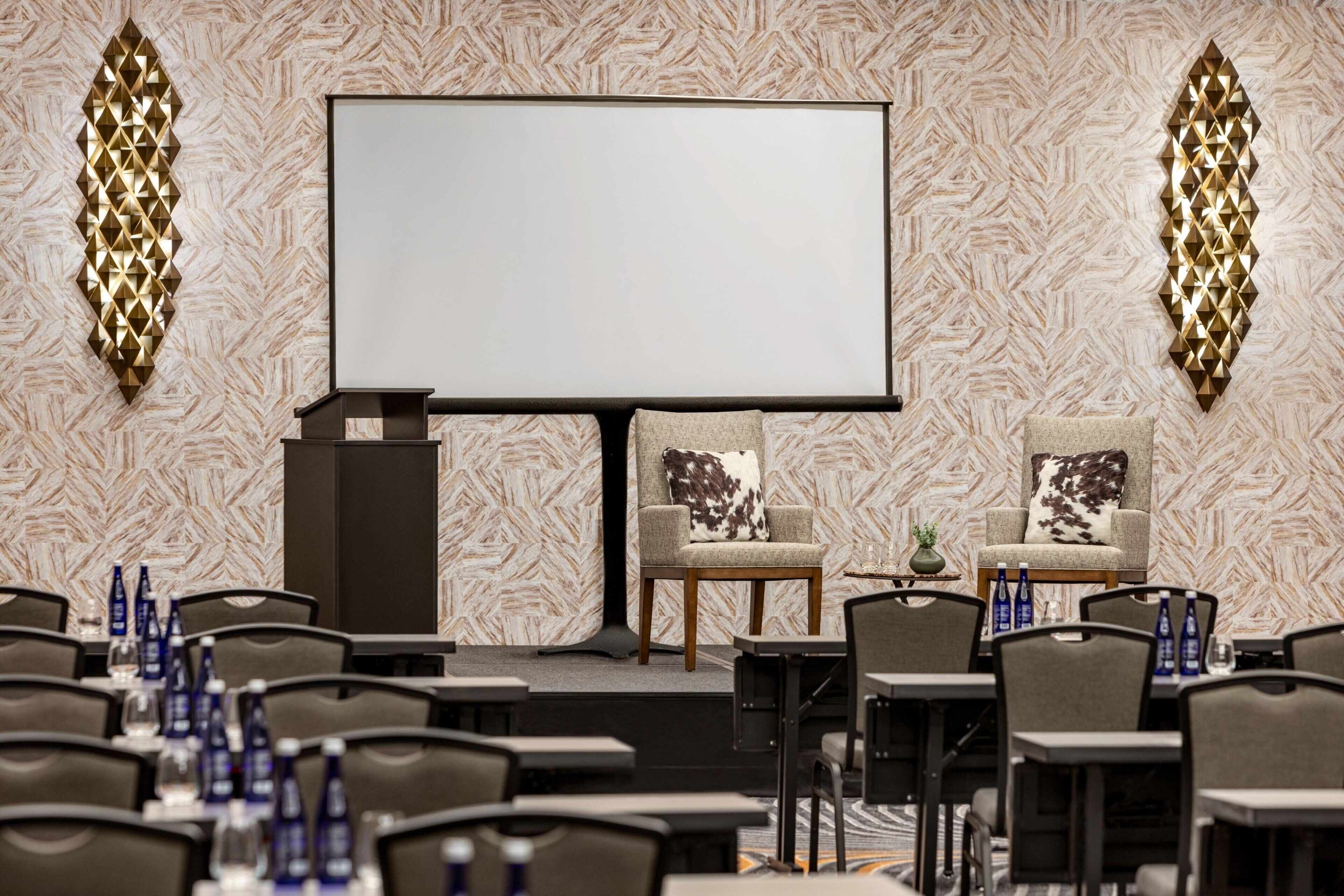 A conference room with a large screen, two chairs with decorative pillows, and tables with water bottles.