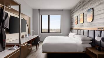 A modern hotel room with a large bed, a desk with a chair, a television, and a view of the city through a large window.
