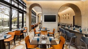 A restaurant with multiple tables and chairs, large windows, and a flat-screen TV on the wall. - Marriott New York JFK Airport (Jamaica)