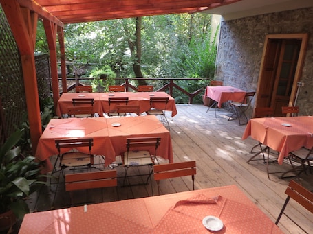 An outdoor patio with multiple tables and chairs, surrounded by greenery and a stone wall.