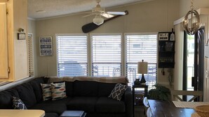 A cozy living room with a dark-colored sofa, a wooden coffee table, and a dining table with chairs. There is a ceiling fan and a chandelier.