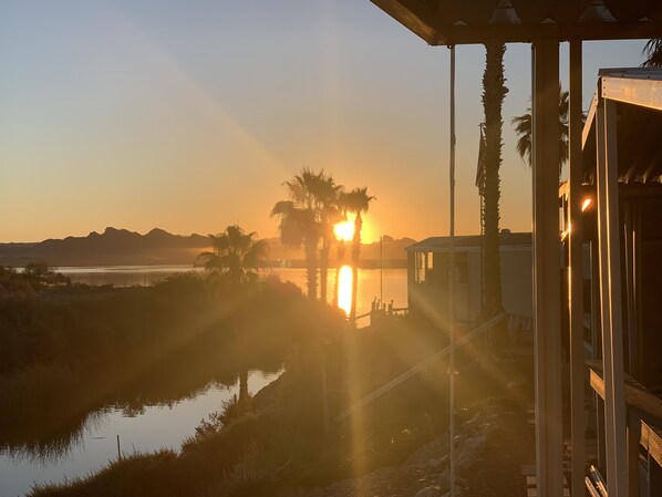 A sunset view from a covered outdoor area with a water body and palm trees in the foreground.