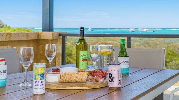 Une table dressée avec une variété de plats et de boissons, notamment une bouteille de vin, une bouteille de gin et une bouteille d’eau gazeuse, le tout disposé devant un paysage marin pittoresque.