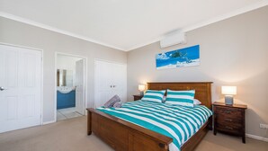 Une chambre à coucher avec un lit en bois, une table de chevet avec une lampe, une salle de bain visible à travers une porte ouverte, et un tableau accroché au mur.