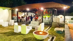 Un área de estar al aire libre con mobiliario blanco, un elemento central de fuego y una estructura cubierta con techo de tejas.