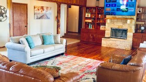 Un salon avec un canapé blanc, des fauteuils en cuir marron, un téléviseur à écran plat, une cheminée en pierre et un lambris en bois.