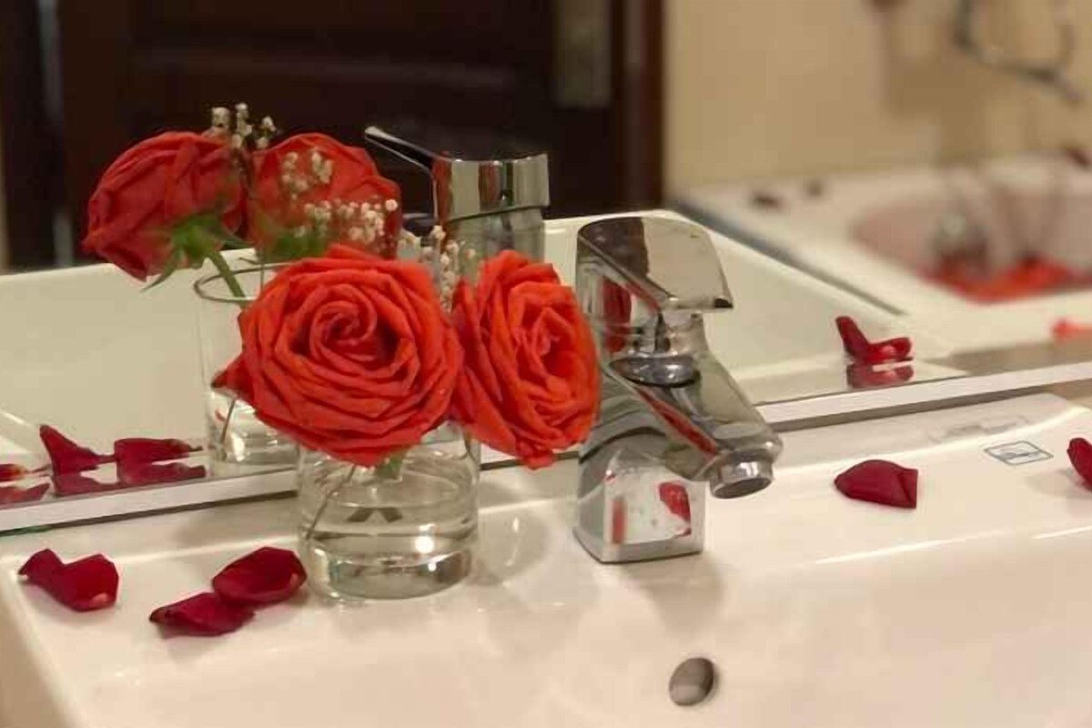 A bathroom sink with a mirror, a vase with red roses, and scattered rose petals.