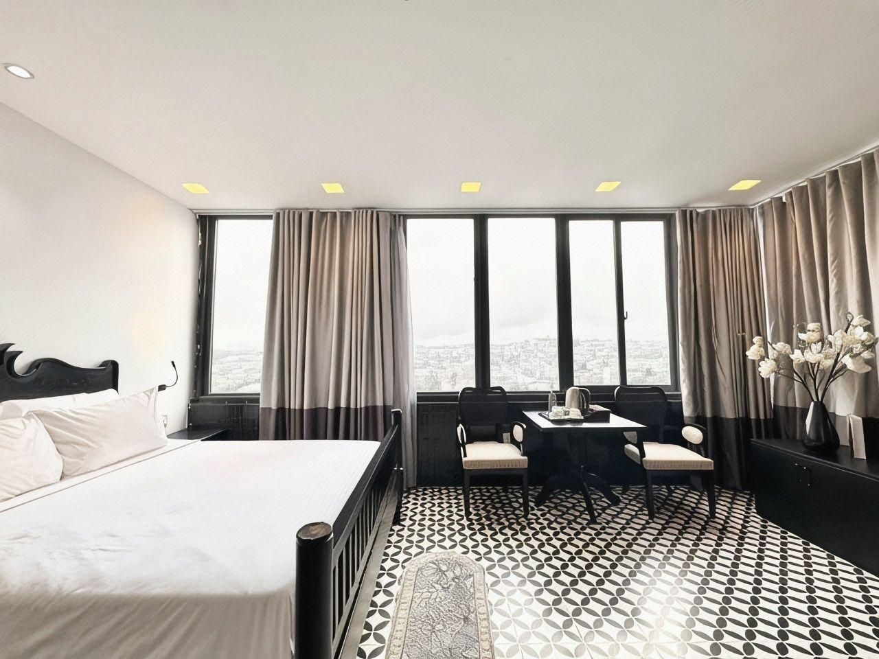Habitación de hotel con una cama grande, un escritorio con silla, y vistas a la ciudad a través de amplias ventanas.