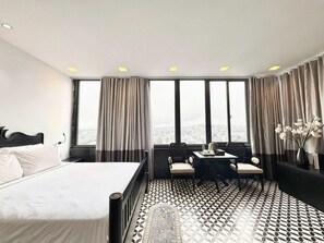 A hotel room with a large bed, a desk with a chair, and a view of the city through large windows.
