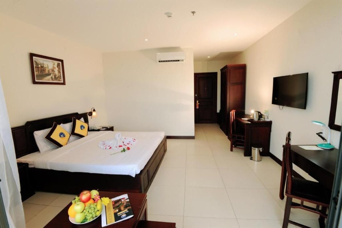 A hotel room with a bed, a desk, a chair, a TV, and a small table with fruit and a book.