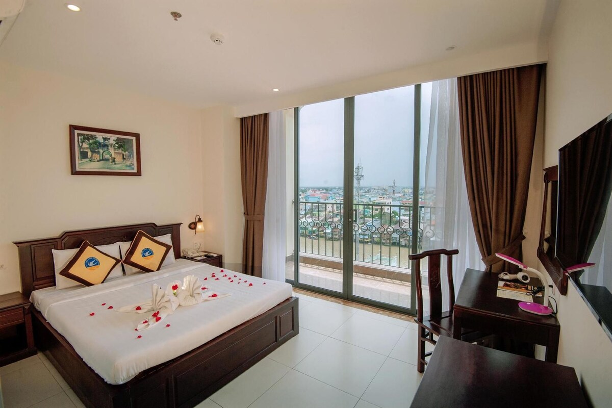 A hotel room with a large bed, a desk, a chair, a window with a view of the city, and a painting on the wall.