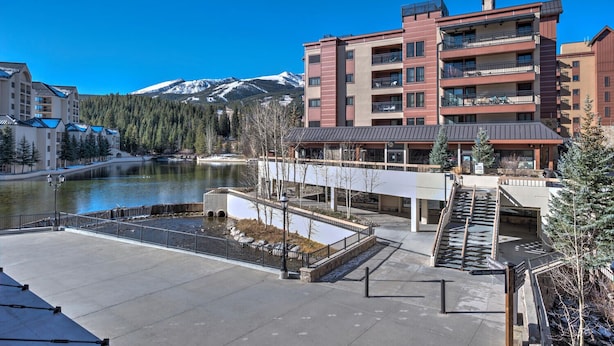 Un hotel moderno con vistas a un lago y montañas. Cuenta con un patio central con una fuente, un corredor cubierto y una escalera que lleva a los pisos superiores.