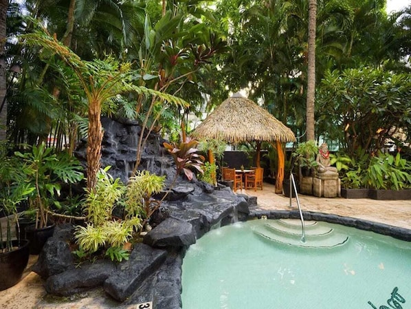 A tropical pool area with a rock formation, a thatched-roof hut, and lush greenery.