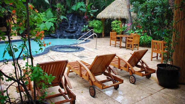 Un área de piscina con sillones de descanso, una sombrilla de paja y exuberante vegetación tropical.