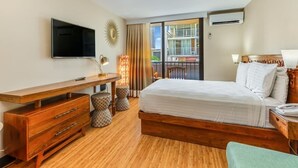 Habitación de hotel con cama de madera, escritorio con televisor, balcón con vista a edificios y aire acondicionado.