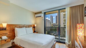 Habitación de hotel con una cama grande, cabecera de madera, mesita de noche con lámpara, ventana con vistas a edificios y balcón con cortinas.