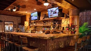 Una zona de bar con una barra de madera rústica, varios taburetes y dos televisores instalados en el techo.