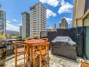 A balcony with a wooden table and chairs, a barbecue grill, and a view of tall buildings.