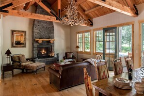 A cozy living room with a fireplace, leather sofa, wooden dining table, and large windows.