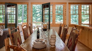 A dining area with a wooden table, chairs, and a bottle of wine. Large windows with a view of trees are visible. - Outpost: Granite Ridge Homestead 3093 - Hot Tub! (Teton Village)