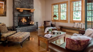 A cozy living room with a stone fireplace, wooden beams, and large windows with a view of trees. - Outpost: Granite Ridge Homestead 3093 - Hot Tub! (Teton Village)