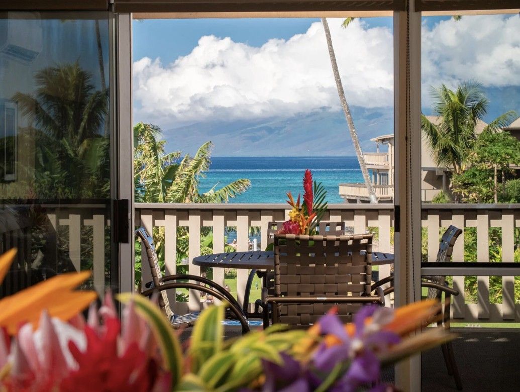 Un balcon avec vue sur l’océan, une table avec des chaises et des plantes tropicales.