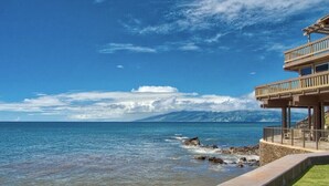 A coastal view with a clear sky, a rocky shoreline, and a building with a balcony overlooking the sea.