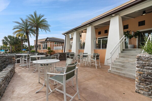 Un espace extérieur aménagé en terrasse, avec des tables et des chaises en métal blanc, un mur en pierre et un escalier menant à un niveau supérieur.