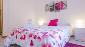 Ett hotellrum med en säng täckt av ett vitt och rosa blommigt överkast, en rosa kudde, en vit handduk och en tavla med en ros som hänger på väggen.