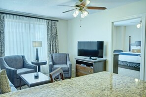 Habitación de hotel con televisor, dos sillones, una mesita, ventilador de techo y una cama.