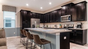 A modern kitchen with dark wood cabinets, a large island with bar stools, stainless steel appliances, and a window with blinds. - Solara Resort 5 Bedroom Townhome Private Pool (Kissimmee)