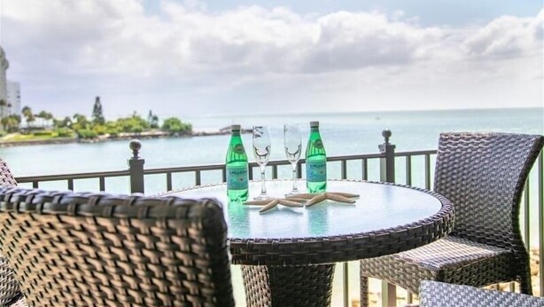Mesa redonda de mimbre con dos botellas y dos vasos en un balcón con vistas a un cuerpo de agua.