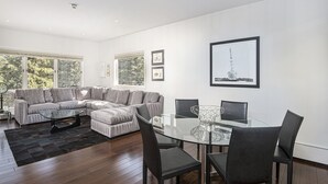 Un salon moderne avec un canapé d’angle, une table à manger en verre et une grande fenêtre donnant sur la forêt.