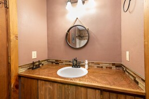 Un baño con tocador de madera, un lavabo blanco, un espejo redondo y una lámpara sobre el espejo.