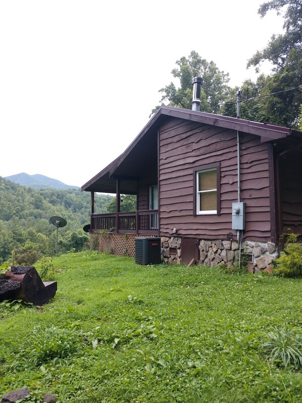 Una cabaña rústica con un porche, rodeada de abundante vegetación y montañas al fondo.