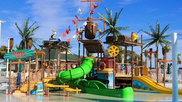 Un parc aquatique avec un toboggan vert, un espace de jeux sur le thème des pirates et des palmiers.