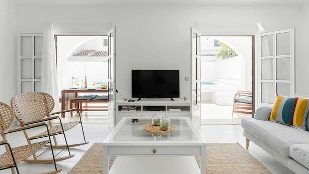 A modern living room with a flat-screen TV on a white coffee table, wicker chairs, a sofa with colorful cushions, and a view through open doors to an outdoor area. - Sea La Vie Quintessential Algarve Villa with Pool in Manta Rota (Vila Nova de Cacela)