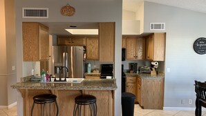 A modern kitchen with wooden cabinets, a stainless steel refrigerator, a granite countertop, and two stools. - Desert Escape with Private Heated Pool & Backyard (Peoria)