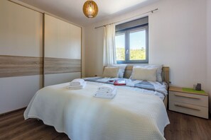 A neatly made bed with a white bedspread, a bedside table with a book and a cup, and a window with curtains. - Villa Legara (Crikvenica)