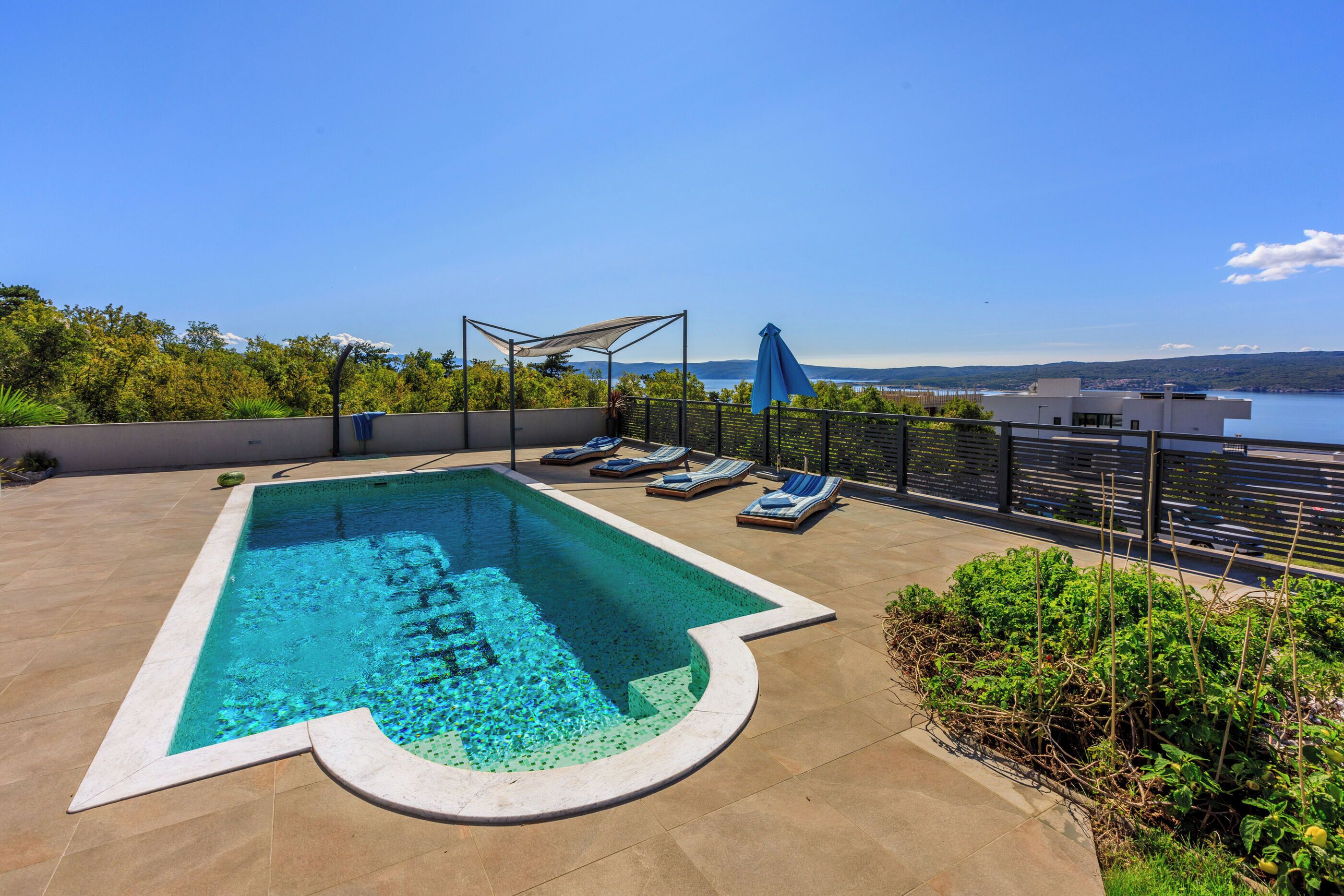 Una piscina con sillas de descanso y una pérgola, rodeada de vegetación y con vistas a un cuerpo de agua.