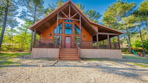 A wooden cabin with a porch, large windows, and a gravel driveway.