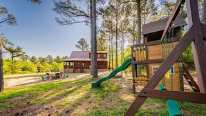 A wooden play structure with a slide, surrounded by trees and a building with a red roof.