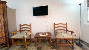 Ein Zimmer mit zwei Holzstühlen, einem kleinen Tisch, einem an der Wand befestigten Fernseher und einer Stehlampe.